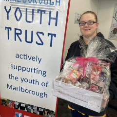 Mackenzie stands to the right of a Marlborough Youth Trust banner. She is holding a gift basket.
