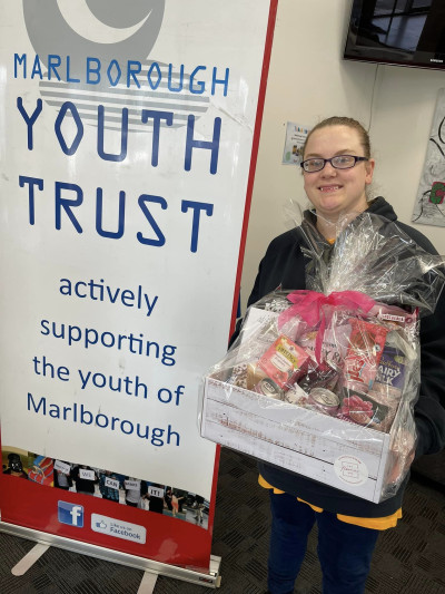Mackenzie stands to the right of a Marlborough Youth Trust banner. She is holding a gift basket.