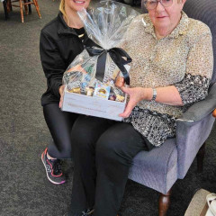 Anne Knight sits in a chair holding a gift basket, with Julie Cadwallender to her left