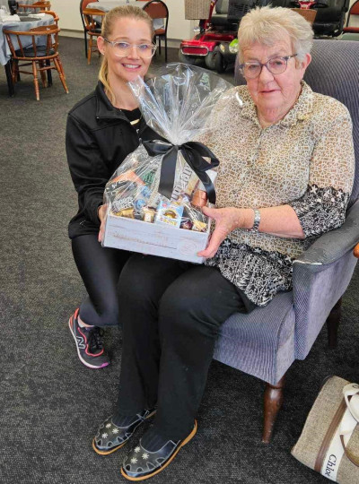 Anne Knight sits in a chair holding a gift basket, with Julie Cadwallender to her left