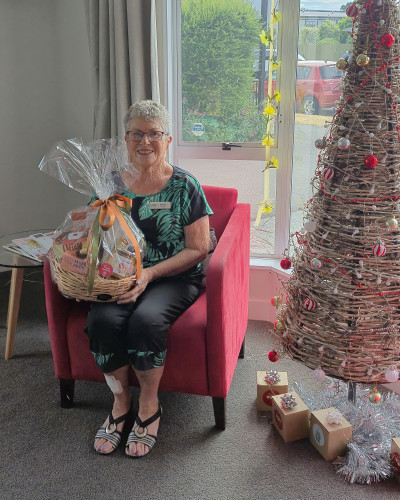 Marrilyn sits in a chair, next to a christmas tree, holding a gift basket