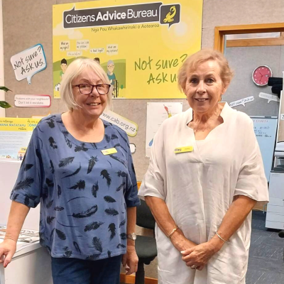 Sue stands on the left in a blue shirt with Jackie on her right in a white shirt. Behind them in the Citizens Advice Bureau sign and office