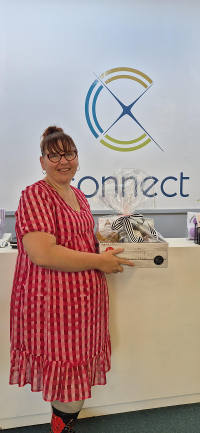 Jess Simmons stands in front of the Connect signage, wearing a red dress, and holding a gift basket